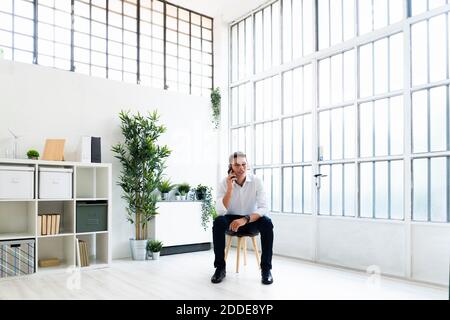 Young businessman talking through mobile phone while sitting on chair in bright creative office Stock Photo