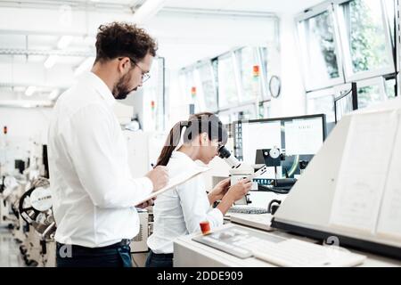 Male technician writing on clipboard while standing by female colleague looking through microscope at laboratory Stock Photo