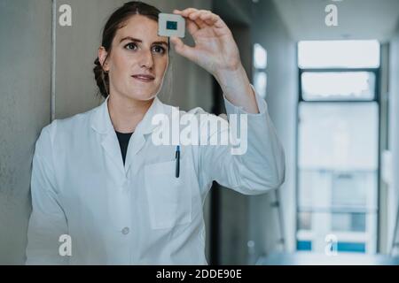 Female scientist holding computer chip while leaning on wall at clinic Stock Photo