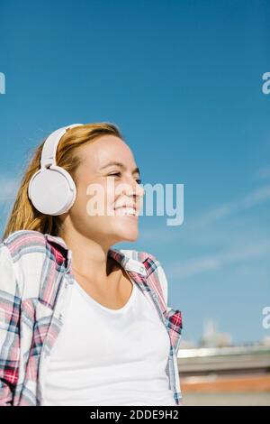 Happy woman listening music through headphone looking away against blue sky on sunny day Stock Photo