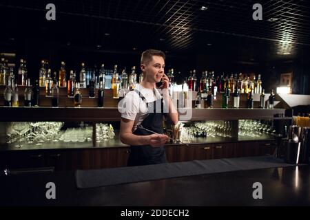The bartender is talking on his smartphone at the bar. Blurry background with bottles. Copy space. Product placement Stock Photo