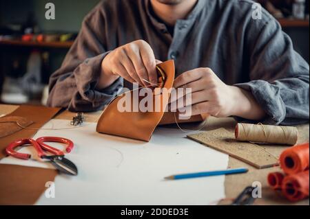 A young shoemaker manually sews decorative elements to leather shoes in the workshop. Stock Photo