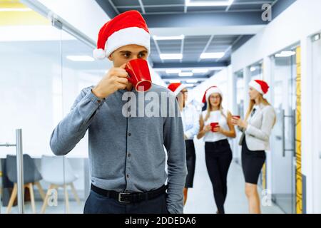 Young businessman wearing Santa Claus hat holding red mug with group of colleagues in the background during coffee break in office. New Year and Chris Stock Photo