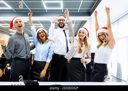 Successful young businessmen in Santa Claus hats, clenching their hands into fists and shouting happily, celebrating the New Year in the office Stock Photo
