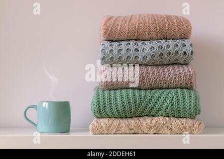 Stack of warm cozy knitted women's sweaters and cup of hot drink on white background. Autumn or winter comfort concept. Stock Photo