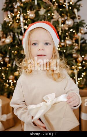 Cute baby girl in Santa Claus hat under Christmas tree with present box. Happy Holidays, New year. Cozy warm winter evening at home. Xmas time