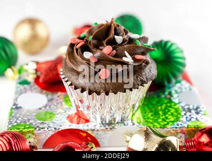 A Christmas Chocolate Cupcake sits on a wrapped foil gift with Christmas ornaments scattered around.  White background. Stock Photo