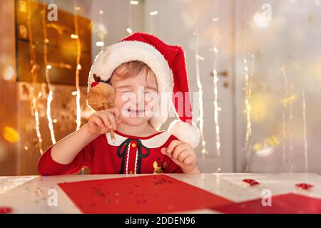 Little happy girl in a red dress and hat writes a letter to Santa Claus. The child makes a wish for Christmas. The baby closed her eyes, writes with a Stock Photo