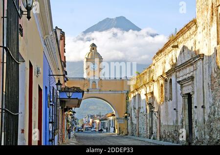 Cloud-shrouded Agua Volcano is one of three volcanoes overlooking the colonial city of Antigua, Guatemala.  Viewed from Calle del Arco, Street of the Arch, the volcanic landmark provides a background for the Santa Catalina Arch, an historic monument built in 1694.  The arch, historically, connected two convents, Convent of the VIrgin and Convent of Santa Catalina.  Nuns were required to not be seen in public thus the arch holds a secret walkway for them to cross between the two convents.  The French Style outdoor clock on the arch was added in the 1800s and needs to be wound every three days. Stock Photo