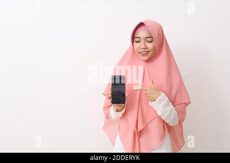 A portrait of happy asian muslim woman wearing a pink veil or hijab and white dress showing and pointing a blank smartphone screen. White background w Stock Photo