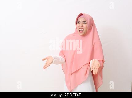 Asian muslim woman in a pink veil or hijab and white dress feels crazy and mad shouting and yelling with aggressive expression and arms raised. Frustr Stock Photo