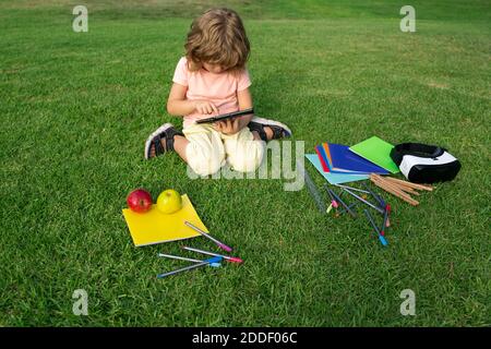 Kids study exam outside. Funny little student boy with tablet, Sit on lawn in park, studying at School backyard. Stock Photo