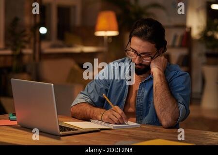 Serious bearded businessman making notes in note pad sitting at his workplace in front of laptop Stock Photo