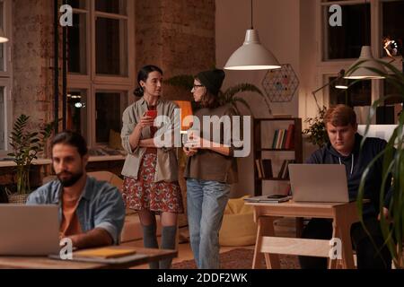 Two young women talking together while standing at office with their colleagues working at their workplaces Stock Photo