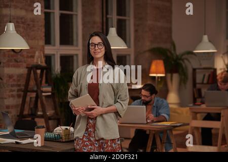 Portrait of young female designer in eyeglasses holding tablet pc and smiling at camera standing at office Stock Photo