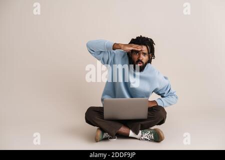 Focused african american guy looking at camera while sitting with laptop on floor isolated over white background Stock Photo