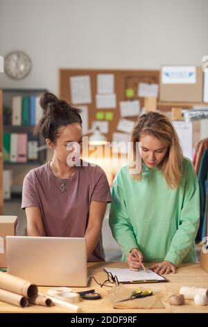 Two young women working in team at the table with computer and documents standing in workshop Stock Photo