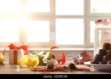 Chocolate truffles in preparation on a kitchen table for the Christmas holidays with ingredients and gift and a window in the background. Front view. Stock Photo