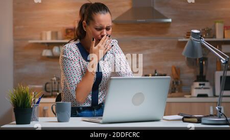 Woman yawning while working remotly from home late at night in the kitchen. Busy exhausted employee using modern technology network wireless doing overtime for job reading typing, searching Stock Photo