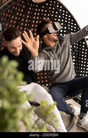 Amused gay couple testing the virtual reality glasses at home. Sitting in the swing on the balcony. High quality photo Stock Photo