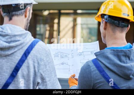 Close up of two builders standing outdoors with an open blueprint discussing the interior of a half completed cottage house. Building, teamwork, partnership concept. Rear view Stock Photo