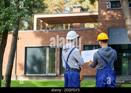 Rear view of two builders standing outdoors with an open blueprint discussing the half completed cottage house. Building, teamwork, partnership concept Stock Photo