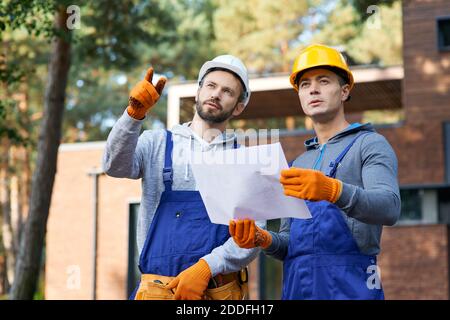 Portrait of two builders looking away while standing outdoors with an open blueprint discussing the half completed cottage house. Building, teamwork, partnership concept Stock Photo
