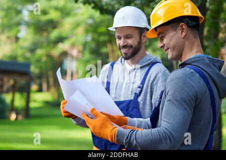 Portrait of two positive builders standing outdoors with an open blueprint discussing the half completed cottage house. Building, teamwork, partnership concept Stock Photo