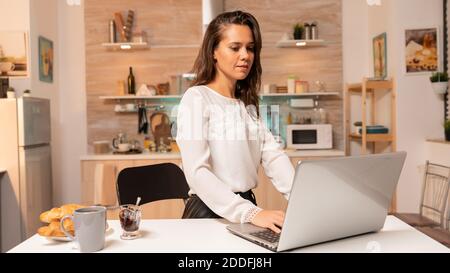 Woman in home kitchen during night hours working on laptop to finish deadline for work. Concentrated entrepreneur in home kitchen using notebook during late hours in the evening. Stock Photo