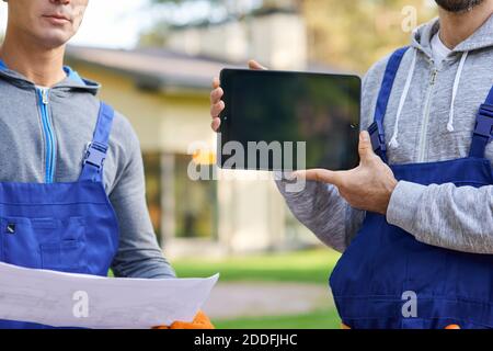 Cropped shot of young male builder showing screen of digital tablet pc with coworker holding blueprint in the background, working at cottage construction site. Selective focus Stock Photo