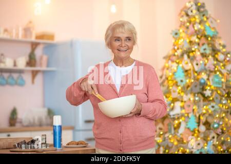 Grey-haired woman in pink shirt stirring something in a bowl and looking cheerful Stock Photo