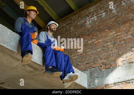 Two cheerful young male builders in blue overalls and hard hats sitting on the concrete floor, taking a break, drinking coffee while working on house construction. Building house concept Stock Photo