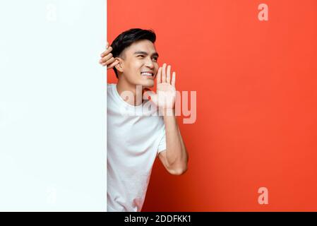 Smiling handsome Asian man about to say something with hand around mouth behind white board on orange background with space for texts Stock Photo
