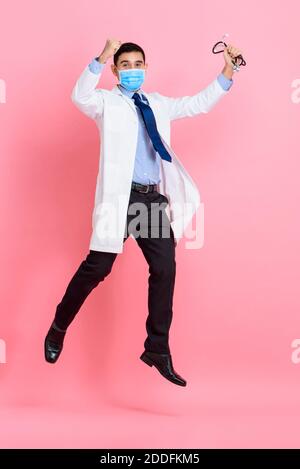 Asian male doctor wearing medical mask raising fists and jumping isolated in pink studio background Stock Photo