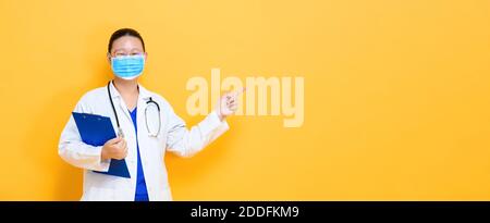 Young Asian woman doctor wearing medical mask pointing hand to empty space aside isolated on yellow banner background Stock Photo