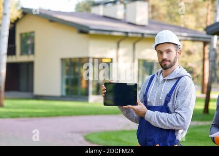 Young male builder smiling at camera, showing digital tablet pc with blank screen while working at cottage construction site. Selective focus Stock Photo
