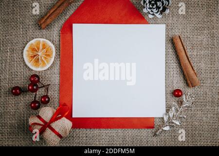 Mockup blank greeting card, red envelope, gift box, cinnamon sticks. Top view flat lay styled Christmas mockup on a rustic rough linen background. Stock Photo