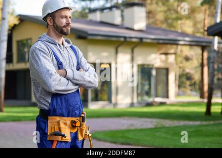 Attractive young male builder in hard hat looking away, posing with arms crossed outdoors while working on cottage construction. Building, people, eco-friendly construction concept Stock Photo