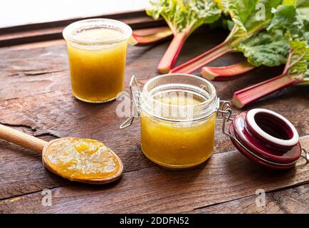 A spoon with homemade rhubarb jam, glass jars of jam and fresh rhubarb stalks on a dark old wooden table near the window, concept of canning food Stock Photo