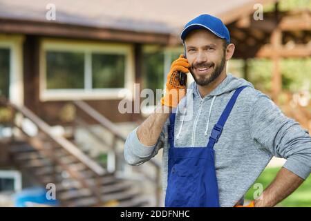 Happy young male builder in uniform smiling at camera while talking on the phone, standing outdoors at cottage construction site. Profession, construction and building concept Stock Photo