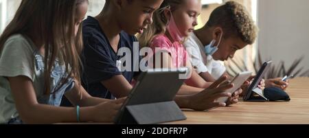 Four pretty kids wear facemasks sit at table use wireless gadgets ignoring each other prefer internet games and virtual communication. Alpha generatio Stock Photo