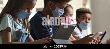Four diverse kids wear facemasks sit at table use wireless gadgets ignoring each other prefer internet games and virtual communication. Alpha generati Stock Photo