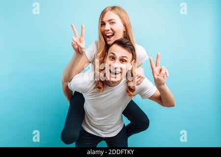 Image of a loving couple, a man and a woman in white T-shirts, happily showing two fingers, a man carrying a joyful woman on his back, on a blue backg Stock Photo