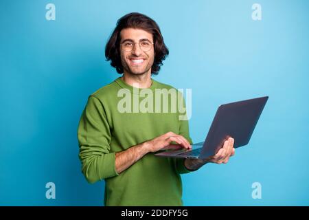 Photo of cheerful programmer guy hold netbook look camera wear glasses green pullover isolated blue color background Stock Photo