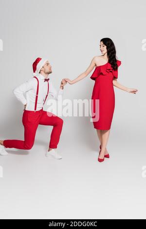 Man in santa hat standing on one knee and holding hand of smiling woman in red dress on grey background Stock Photo