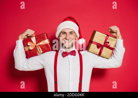 Cheerful man in santa hat holding gift boxes on red background Stock Photo