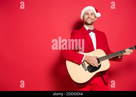 Smiling man in santa hat playing acoustic guitar on red background Stock Photo