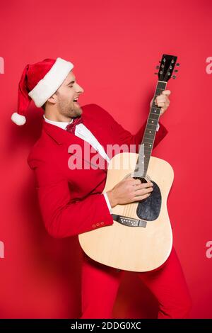 Positive man in santa hat playing acoustic guitar on red background Stock Photo