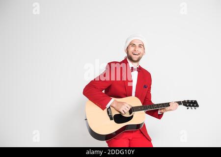 Young man in suit and santa hat smiling at camera while playing acoustic guitar on grey background Stock Photo