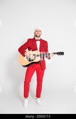 Positive man in suit and santa hat playing acoustic guitar on grey background Stock Photo